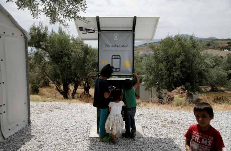 This is a photo of Children stand around a solar powered charging station for mobile devices designed by a group of university students, at the municipality-run camp of Kara Tepe on the island of Lesbos, Greece, June 14, 2016.